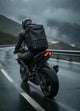 Motorcyclist riding on a wet mountain road wearing the LUMA Control backpack, rear view