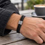 Man wearing a black leather bracelet with two engraved beads on his wrist at a cafe table