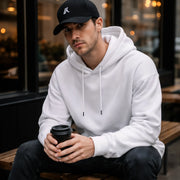 Man wearing the LUMA White Velvet oversized hoodie and cap, sitting at a cafe holding coffee