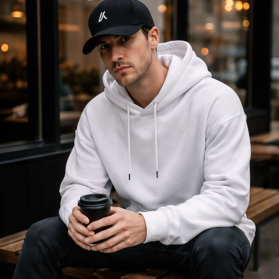 Man wearing the LUMA White Velvet oversized hoodie and cap, sitting at a cafe holding coffee